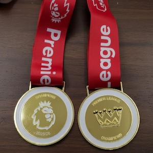 以下が含まれることがあります： 白い縁取りが施された2つの金色の円形メダル。それぞれ「Premier League」の白い文字が入った赤いリボンが付いています。片方のメダルにはライオンの頭のロゴと「2025/26」が、もう片方には王冠のグラフィックと「Premier League Champions」が刻まれています。