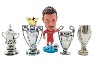 Könnte beinhalten: Eine silberne Trophäe mit einer goldenen Krone obenauf, eine silberne Trophäe mit der Aufschrift "Premier League", eine silberne Trophäe mit der Aufschrift "UEFA Champions League", eine silberne Trophäe mit einem runden Sockel und eine Figur eines Fußballspielers in einem roten Trikot.