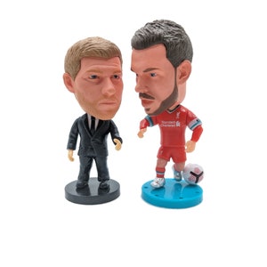 K&ouml;nnte beinhalten: Zwei Bobblehead-Figuren, eine von einem Mann im schwarzen Anzug und die andere von einem Mann im roten Fu&szlig;balltrikot mit dem Text "Standard Chartered" auf der Vorderseite. Der Fu&szlig;ballspieler h&auml;lt einen Fu&szlig;ball.