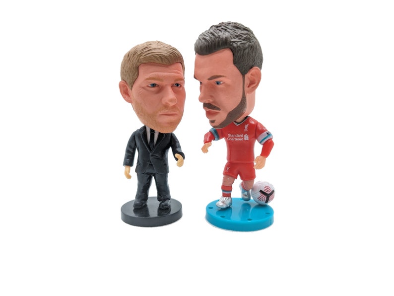 K&ouml;nnte beinhalten: Zwei Bobblehead-Figuren, eine von einem Mann im schwarzen Anzug und die andere von einem Mann im roten Fu&szlig;balltrikot mit dem Text "Standard Chartered" auf der Vorderseite. Der Fu&szlig;ballspieler h&auml;lt einen Fu&szlig;ball.