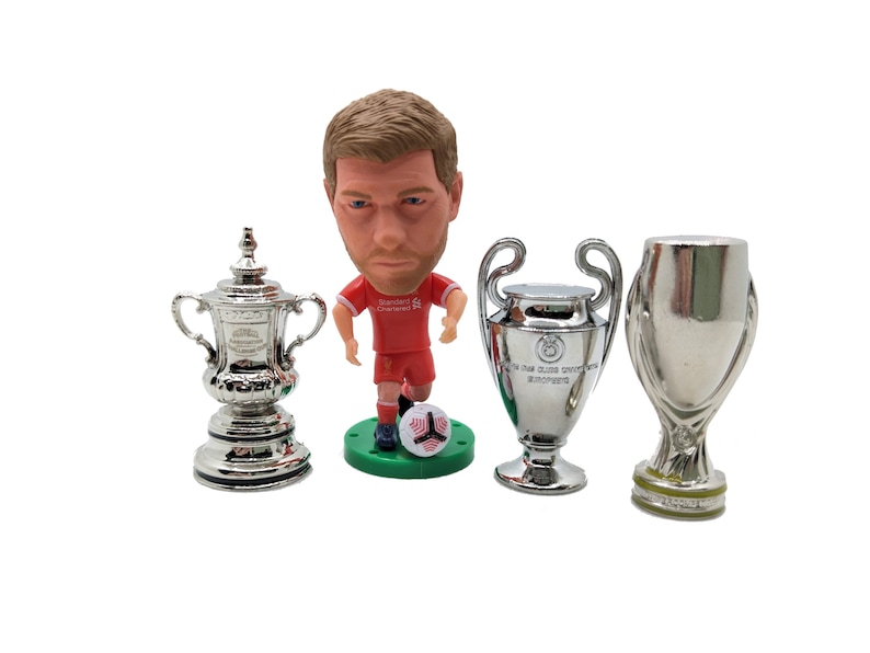 K&ouml;nnte beinhalten: Eine silberne Miniatur-Replik des FA Cups, eine silberne Miniatur-Replik der UEFA Champions League Troph&auml;e und eine silberne Miniatur-Replik der UEFA Europa League Troph&auml;e, zusammen mit einer Figur eines Fu&szlig;ballspielers in einem roten Trikot.
