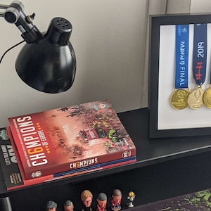 Könnte beinhalten: Ein schwarzer, gerahmter Vitrinenständer mit fünf Medaillen und Bändern. Die Bänder sind blau, rot und weiß mit dem Text "Premier League", "FIFA", "2019", "FIFA", und "Madrid Final". Die Medaillen sind aus Gold und Silber.