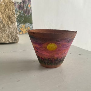 May include: A small terracotta pot with a painted sunset scene. The pot is painted with a pink, purple, and orange sunset with a yellow sun in the center. The pot is perfect for small plants or succulents.
