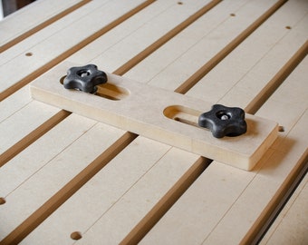 May include: A wooden workbench with a sliding wooden clamp with two black knobs. The clamp is positioned on the workbench surface.