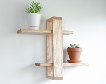 May include: A wooden wall shelf with two shelves and a vertical support. The shelf is made of light wood and has a modern design. Two small potted plants are on the shelves.