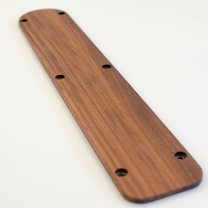 Puede incluir: Una manilla de puerta larga y rectangular de madera con la parte superior redondeada y un acabado de veta de madera natural. La manilla tiene cinco agujeros perforados uniformemente espaciados.