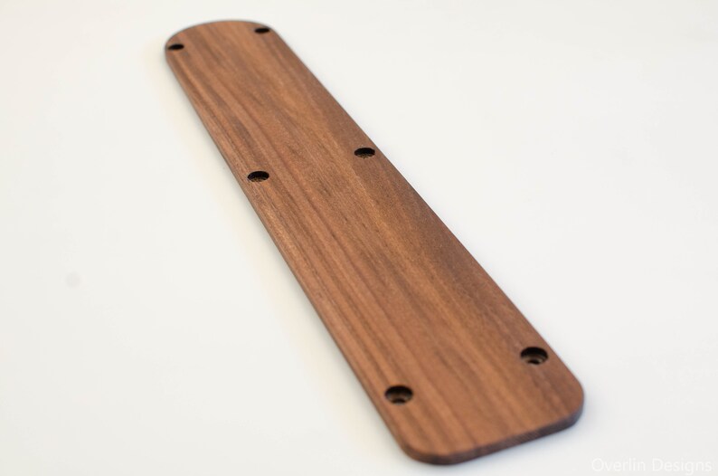 Puede incluir: Una manilla de puerta larga y rectangular de madera con la parte superior redondeada y un acabado de veta de madera natural. La manilla tiene cinco agujeros perforados uniformemente espaciados.