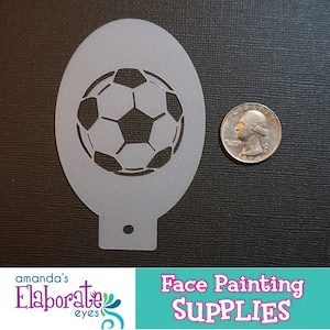 May include: A light gray soccer ball stencil with a quarter for size comparison. The stencil is oval-shaped with a soccer ball design cut out in the center. The bottom of the stencil has a small tab with a hole. The image also includes the text "Face Painting SUPPLIES".