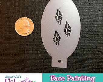 May include: A silver face painting stencil with three diamond shapes, next to a quarter. The stencil is oval-shaped with a small hole at the bottom. The image also includes the text "Face Painting SUPPLIES".