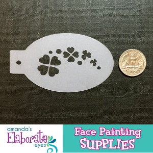 Puede incluir: Un esténcil blanco de plástico para pintar caras con un diseño de trébol. El esténcil tiene forma ovalada con una pequeña pestaña y un agujero. Una moneda está al lado del esténcil para comparar el tamaño. El texto "Face Painting SUPPLIES" está en la parte inferior.