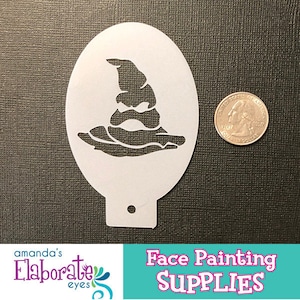 May include: A white plastic face painting stencil featuring a wizard's hat design. The stencil is oval-shaped with a small hole at the bottom. A U.S. quarter coin is next to the stencil for size comparison. The image also includes the text "Face Painting SUPPLIES".