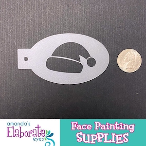 Puede incluir: Un esténcil plateado de gorro de Papá Noel para pintar caras, con una moneda para comparar el tamaño. El esténcil tiene forma ovalada con una pequeña pestaña y un agujero en un extremo. Las palabras "Face Painting SUPPLIES" están en la esquina inferior derecha.