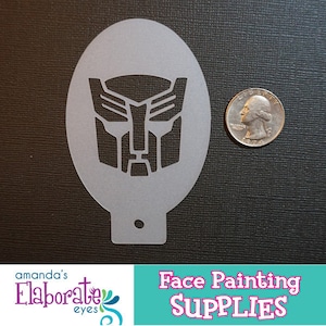 Puede incluir: Un esténcil de plástico gris con el símbolo Autobot, utilizado para pintar caras. El esténcil tiene forma ovalada con un pequeño agujero en la parte inferior. Una moneda de veinticinco centavos estadounidense está al lado del esténcil para comparar el tamaño. También incluye el texto "Face Painting SUPPLIES".