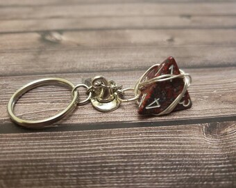May include: A keychain featuring a red and black polyhedral die, wrapped in silver wire. The die is attached to a silver ring and a small charm. The background is a wooden surface.