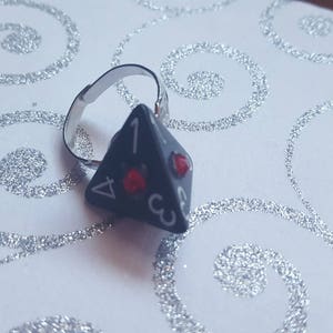May include: A black and silver ring with a four-sided dice charm. The dice has red gems on each side and the numbers 1, 2, 3, and 4 are printed on the sides.