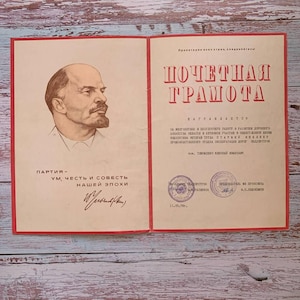 May include: A vintage Soviet certificate of honor featuring a portrait of Vladimir Lenin on the left side and the red Cyrillic text POCHETNAYA GRAMOTA on the right. The document includes official stamps, a signature, and printed Russian text.