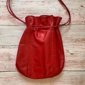 Könnte beinhalten: Eine leuchtend rote Kordelzugtasche aus Leder mit einem weichen, unstrukturierten Körper, vertikalen Steppnähten und einem dünnen, passenden roten Schulterriemen aus Leder, der die obere Öffnung beim Zuziehen sicher verschließt.