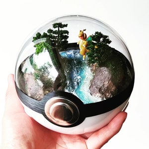 May include: A glass dome terrarium with a miniature landscape featuring a small waterfall, green plants, and a small figurine of a Pokemon character. The terrarium is shaped like a Pokeball, with a black band and a red button.