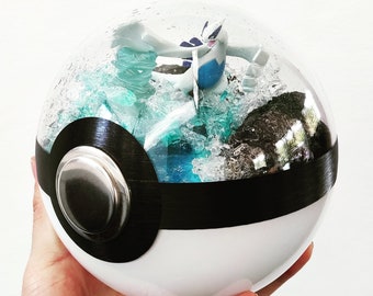 May include: A white and black Poké Ball with a clear dome, featuring a detailed diorama of a Pokémon character amidst blue and turquoise water-like elements. The diorama includes a white and blue Pokémon figure and a dark rock formation.