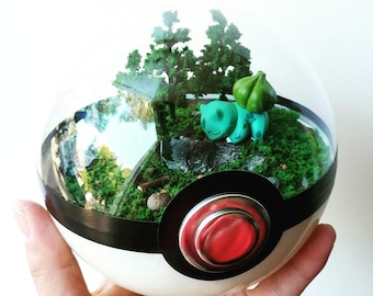 Puede incluir: Un recipiente esférico transparente diseñado como una Poké Ball con un exterior rojo y blanco, que alberga un diorama en miniatura con una pequeña criatura verde con un bulbo en la espalda, árboles verde oscuro y suelo cubierto de musgo.