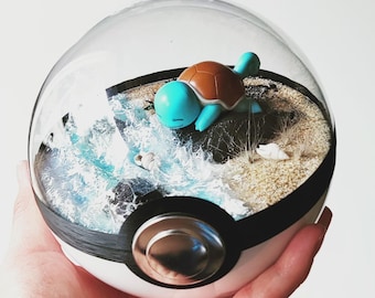 Puede incluir: Un recipiente esférico transparente diseñado como una Poké Ball que contiene una figura de tortuga azul claro sobre una roca oscura. El interior incluye una zona de playa arenosa, pequeñas conchas blancas y agua azul texturizada.