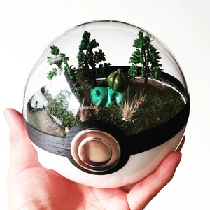 Könnte beinhalten: Ein Glaskuppel-Terrarium mit einer schwarz-weißen Basis, das einem Pokéball ähnelt, mit einer kleinen grünen Figur und Miniaturpflanzen.