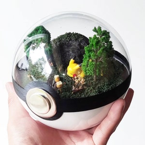 May include: A glass dome terrarium shaped like a Pokeball with a small yellow Pikachu figurine inside. The terrarium is filled with green moss, small trees, and a black rock.