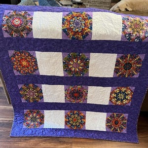 May include: A handmade quilt with a purple border and backing. The quilt features colorful, circular designs in the center, alternating with white rectangular blocks. The circular designs have intricate patterns in shades of red, orange, blue, and green.