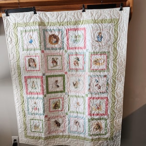 May include: A white quilt with a repeating pattern of square blocks featuring illustrations of animals in pastel colors. The blocks are framed in pink and green, with a green border and decorative stitching.