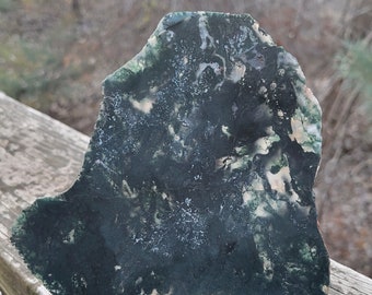 May include: A large, irregularly shaped piece of green moss agate with white and light green markings. The agate is sitting on a wooden surface.