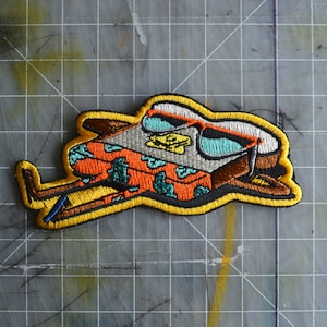 Op de afbeelding: Geborduurde patch met een gestileerd ontwerp van een voertuig met een zonnebril. De patch heeft een gele rand en bevat oranje, bruine, teal en gele kleuren. Het voertuig heeft een patroon en draagt een zonnebril.