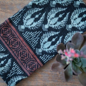 May include: A black and grey knitted scarf with a geometric pattern and a brown and red border. The scarf is folded and lying on a wooden surface.
