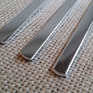 May include: Three silver metal strips, each with a rounded edge, are laid out on a beige woven fabric background.