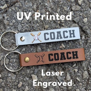 Könnte beinhalten: Zwei rechteckige Schlüsselanhänger mit Metallringen. Der obere Schlüsselanhänger ist hellgrau mit einer UV-gedruckten Baseball- und Schlägergrafik und dem Wort COACH. Der untere Schlüsselanhänger ist braun mit einer Lasergravur.