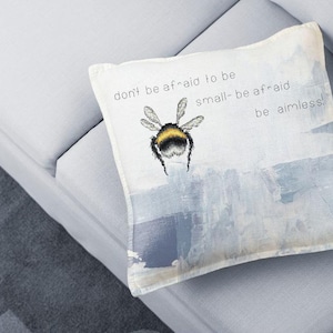 Puede incluir: Cojín cuadrado con una ilustración de abeja y el texto "don't be afraid to be small-be afraid be aimless!". El cojín está sobre un sofá gris claro. El diseño tiene un fondo blanco con toques de azul y gris.