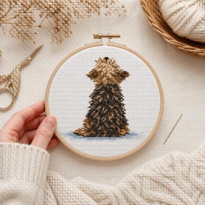 May include: A cross-stitch embroidery of a dog in a wooden hoop. The dog is brown and black, with its head tilted upwards. The background is white, and the hoop is held by a person wearing a cream-colored sweater. A pair of gold scissors and a basket of yarn are in the background.