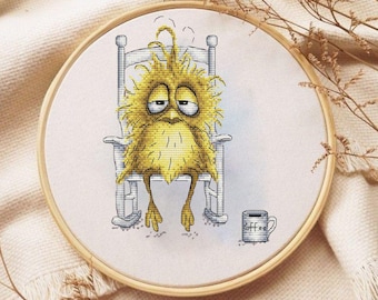 May include: Cross-stitch embroidery in a wooden hoop depicts a sleepy, yellow creature with droopy eyes sitting in a rocking chair. A small coffee mug sits nearby. The design is on white fabric.