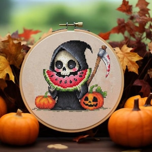 May include: A cross-stitch design in a wooden hoop features a cartoon Grim Reaper holding watermelon. Two orange pumpkins and a jack-o'-lantern are in front of the hoop. The background includes fall leaves and pumpkins.