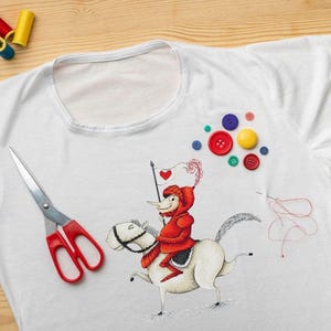 May include: A white t-shirt with a red and white illustration of a knight riding a horse. The illustration is surrounded by colorful buttons and spools of thread.