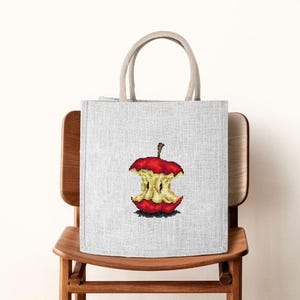 May include: A light gray tote bag with a detailed embroidered image of an apple core. The apple core is red and yellow, with a brown stem. The bag has beige handles and is sitting on a wooden chair.