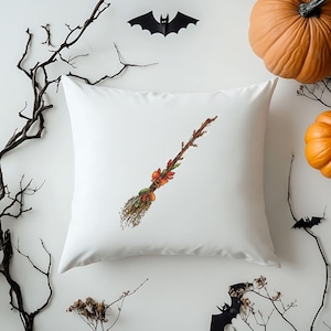 May include: White square pillow with a decorative embroidered broom design. The broom features orange, green, and brown accents, with a bow and pumpkins. Halloween-themed decor.