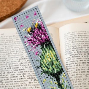 Op de afbeelding: Kruissteek bladwijzer met een bij en distel ontwerp. De bladwijzer heeft een grijze achtergrond met een gedetailleerde afbeelding van een bij en paarse distelbloemen. De bladwijzer is rechthoekig en lijkt handgemaakt.