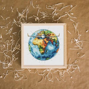 Puede incluir: Una obra de arte en punto de cruz enmarcada que representa la Tierra con un avión de pasajeros que la atraviesa. El globo está representado en tonos azules, verdes, naranjas y marrones. El avión es blanco y gris. El marco es de madera clara.