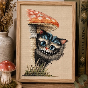 May include: A framed cross-stitch embroidery on beige fabric featuring a black cat with blue eyes and a wide, toothy grin peeking from behind a large red and white spotted mushroom. The cat has blue tufts of fur on its head and ears.