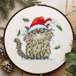 May include: Cross-stitch embroidery of a grumpy cat wearing a Santa hat. The cat is gray and white with an angry expression. Green holly sprigs with red bows are scattered around the cat. The embroidery is in a wooden hoop.