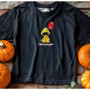 May include: A black t-shirt with a yellow raincoat character holding a red balloon. The character is standing on a white background. The t-shirt is surrounded by pumpkins.