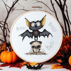 May include: Cross-stitch embroidery in a wooden hoop featuring a cartoon bat with large eyes, standing on a skull. The bat has black wings and yellow ears. The word "BOO!" is written on the white fabric. Halloween theme.