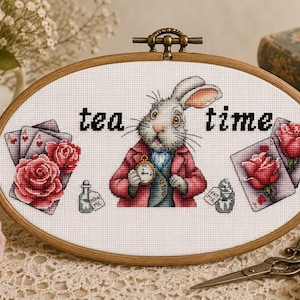 May include: An oval wooden embroidery hoop containing a cross-stitch design of a rabbit wearing a red coat and holding a pocket watch. The text "tea time" is stitched above the rabbit, flanked by playing cards, red roses, and small bottles.