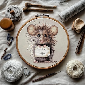 Puede incluir: Una bordado de punto de cruz de un ratón marrón y blanco en un aro de madera. El ratón sostiene un cartel que dice "Necesito mi café..."