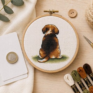May include: A circular wooden embroidery hoop containing a cross-stitch design of a brown, black, and white beagle dog viewed from behind, sitting on a small patch of green grass against a white fabric background, with a one-euro coin for scale.